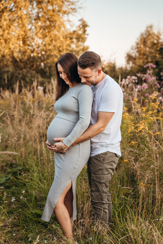 Schwangere Frau posiert mit ihrem Partner, beide lächeln glücklich und halten den Babybauch während einer Fotosession im Wald